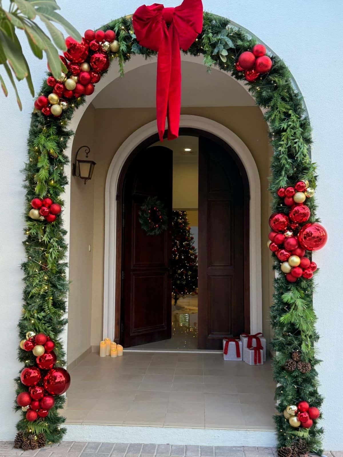 Arched villa entrance framed with an evergreen garland and clusters of red and gold baubles, crowned with a large red bow