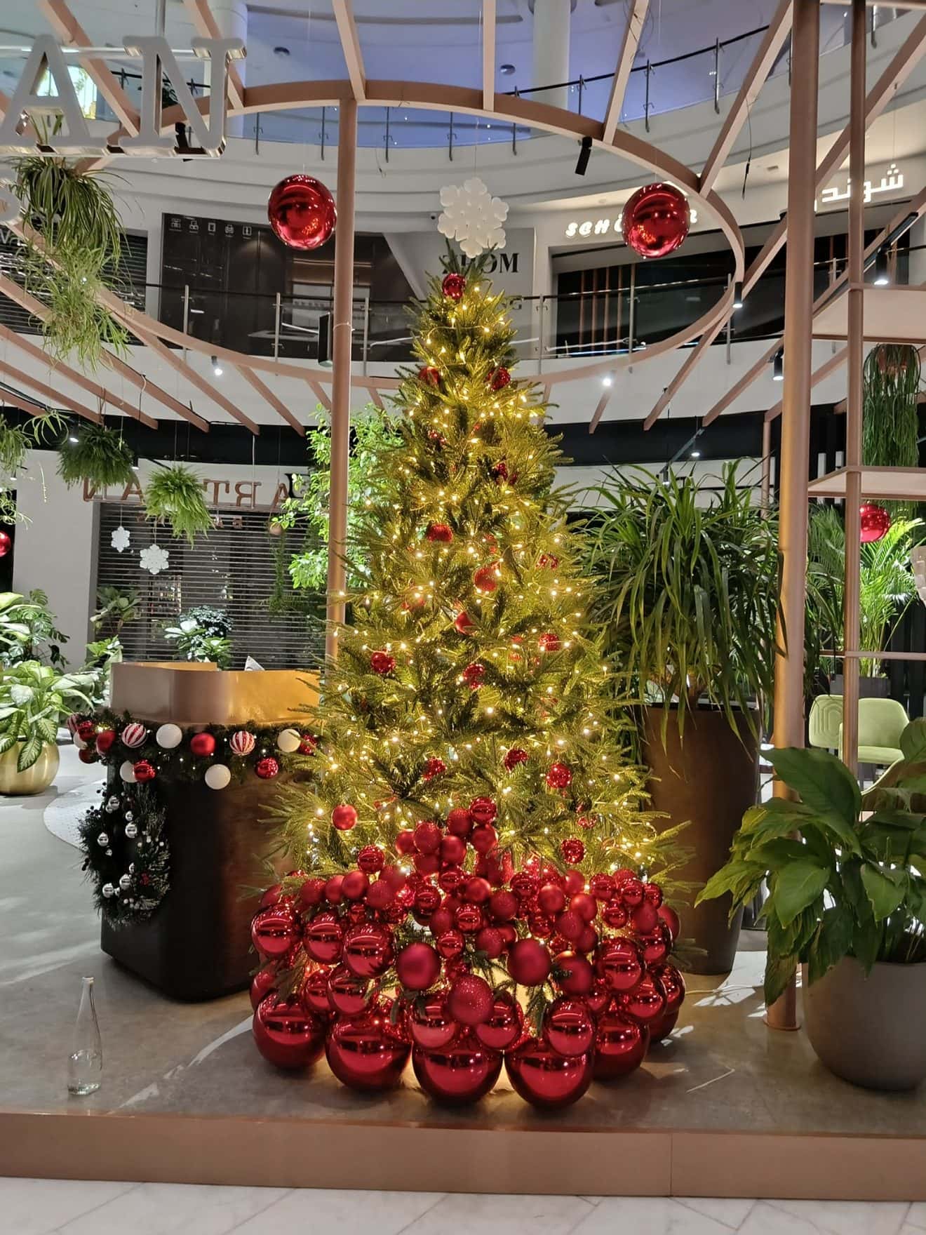 Christmas tree glowing with warm gold lights, its base heaped with red baubles, in a double-height atrium with plants