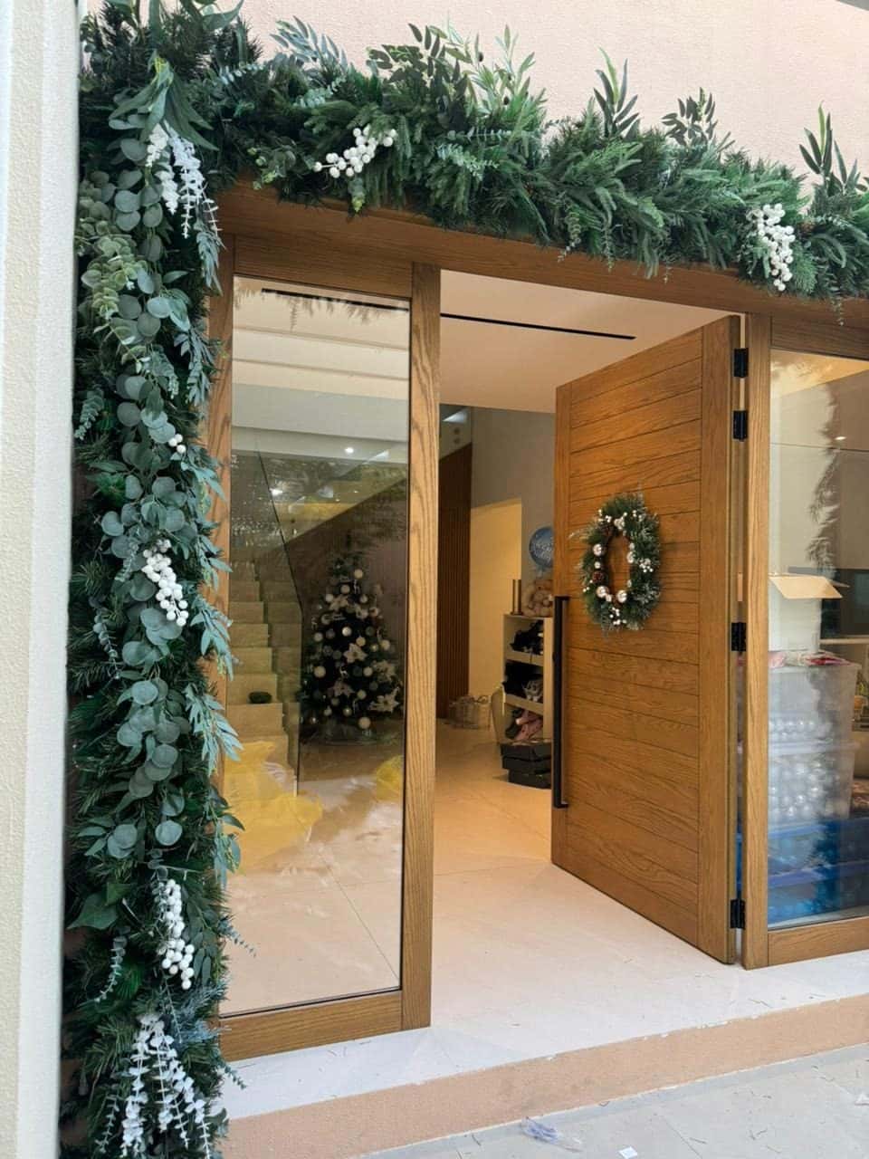 Soft greenery garland with white sprays framing a glass-fronted entrance, with a wreath on the open wooden door