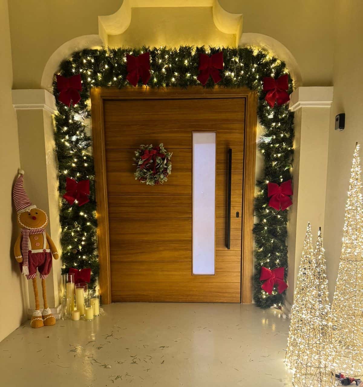Wooden front door framed by a lit garland with red bows, with lit wire cone trees and candle-style lights beside it in the evening