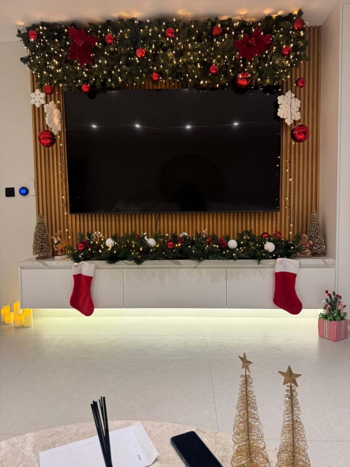 Lit garland with red poinsettias and baubles framing a television wall, with two red stockings hanging below and small cone trees on the console