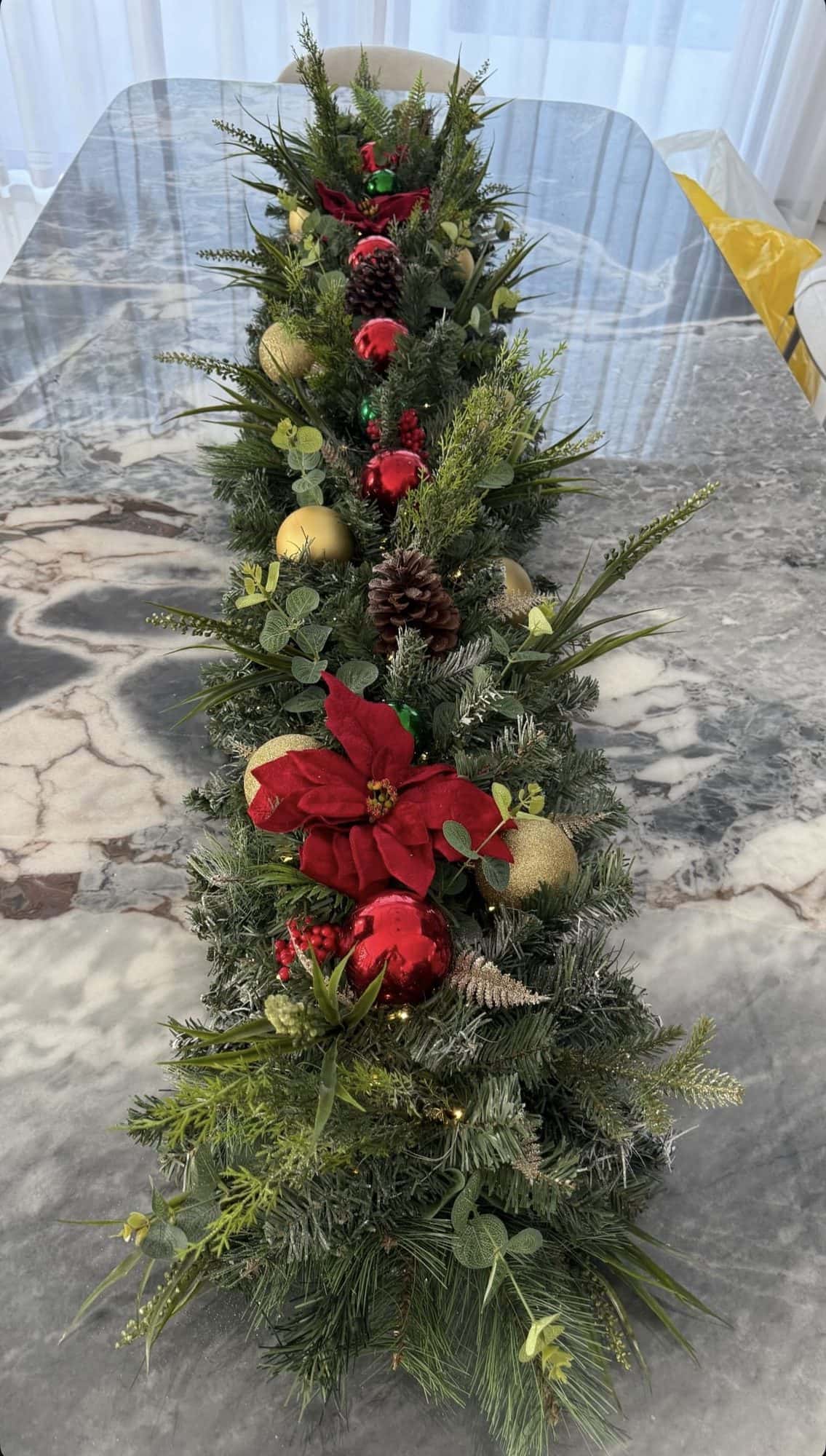 Long evergreen table runner with a red poinsettia, gold baubles and pine cones on a grey marble dining table