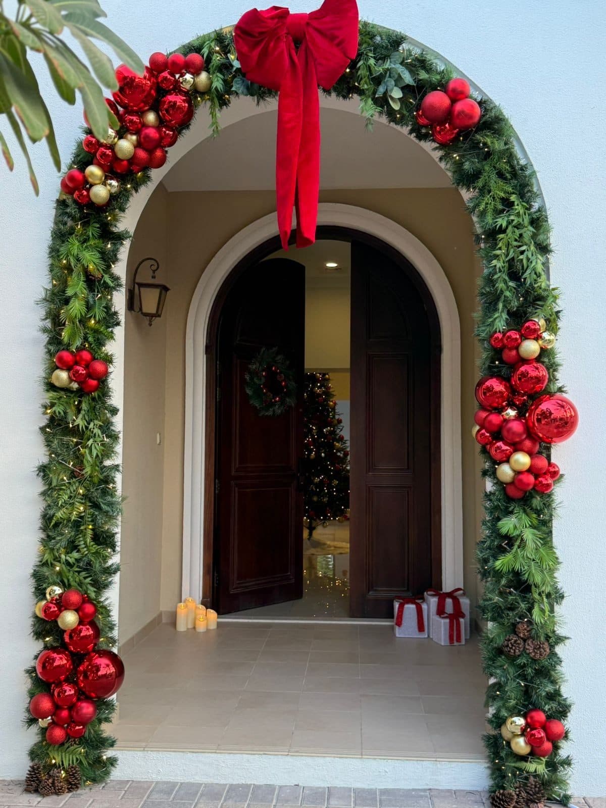 Arched villa entrance framed with an evergreen garland and clusters of red and gold baubles, crowned with a large red bow