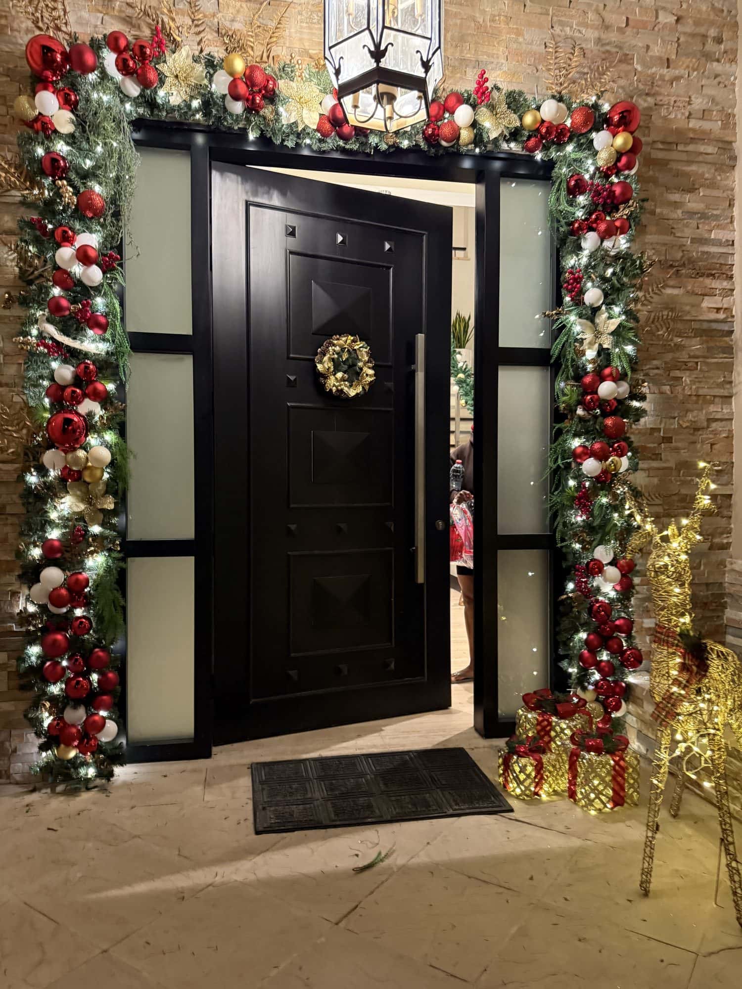 Black pivot front door at night framed by an evergreen arch of red, white and gold baubles, with a gold wreath, wrapped gift boxes and a lit gold reindeer beside it