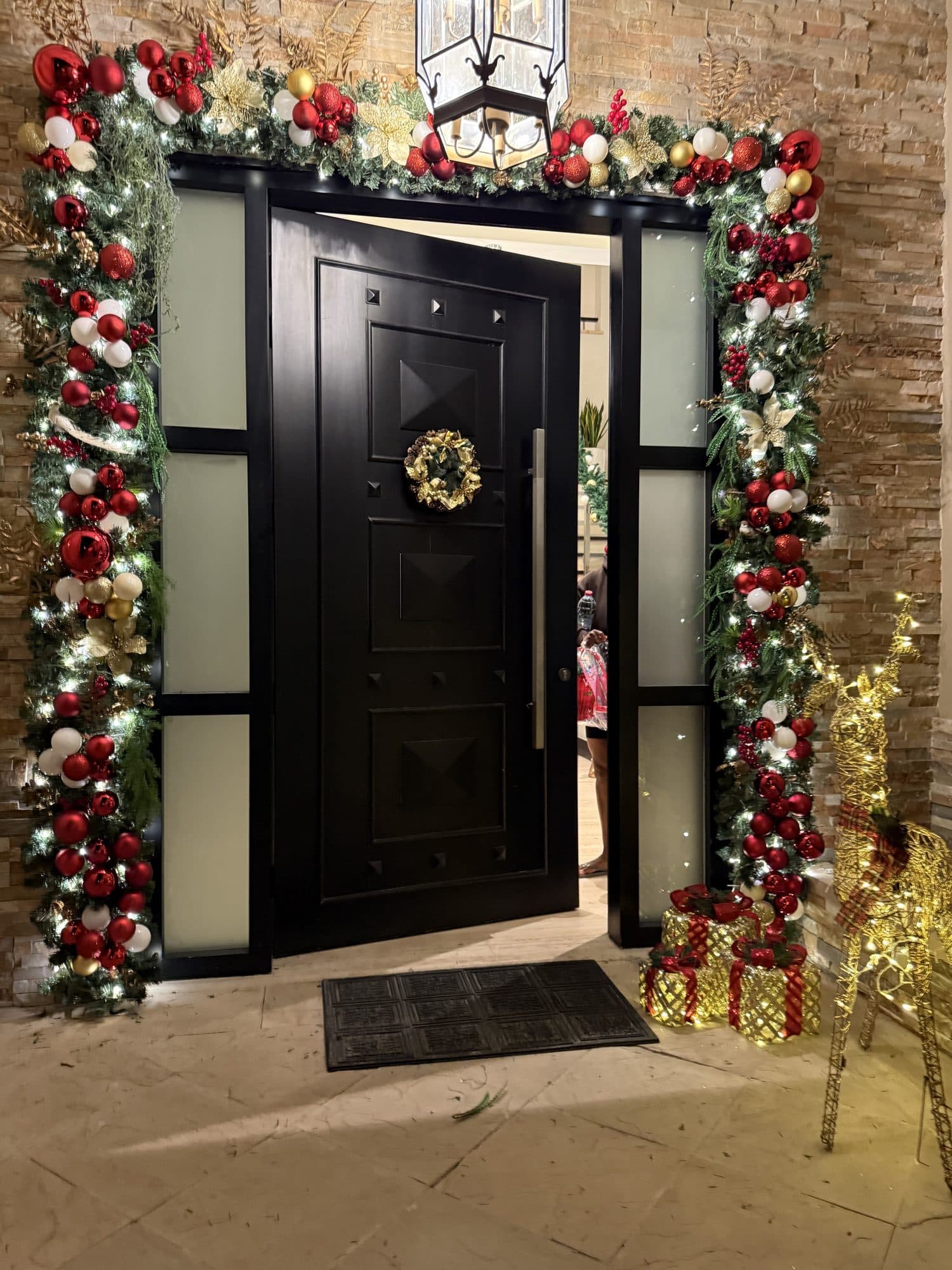 Black pivot front door at night framed by an evergreen arch of red, white and gold baubles, with a gold wreath, wrapped gift boxes and a lit gold reindeer beside it