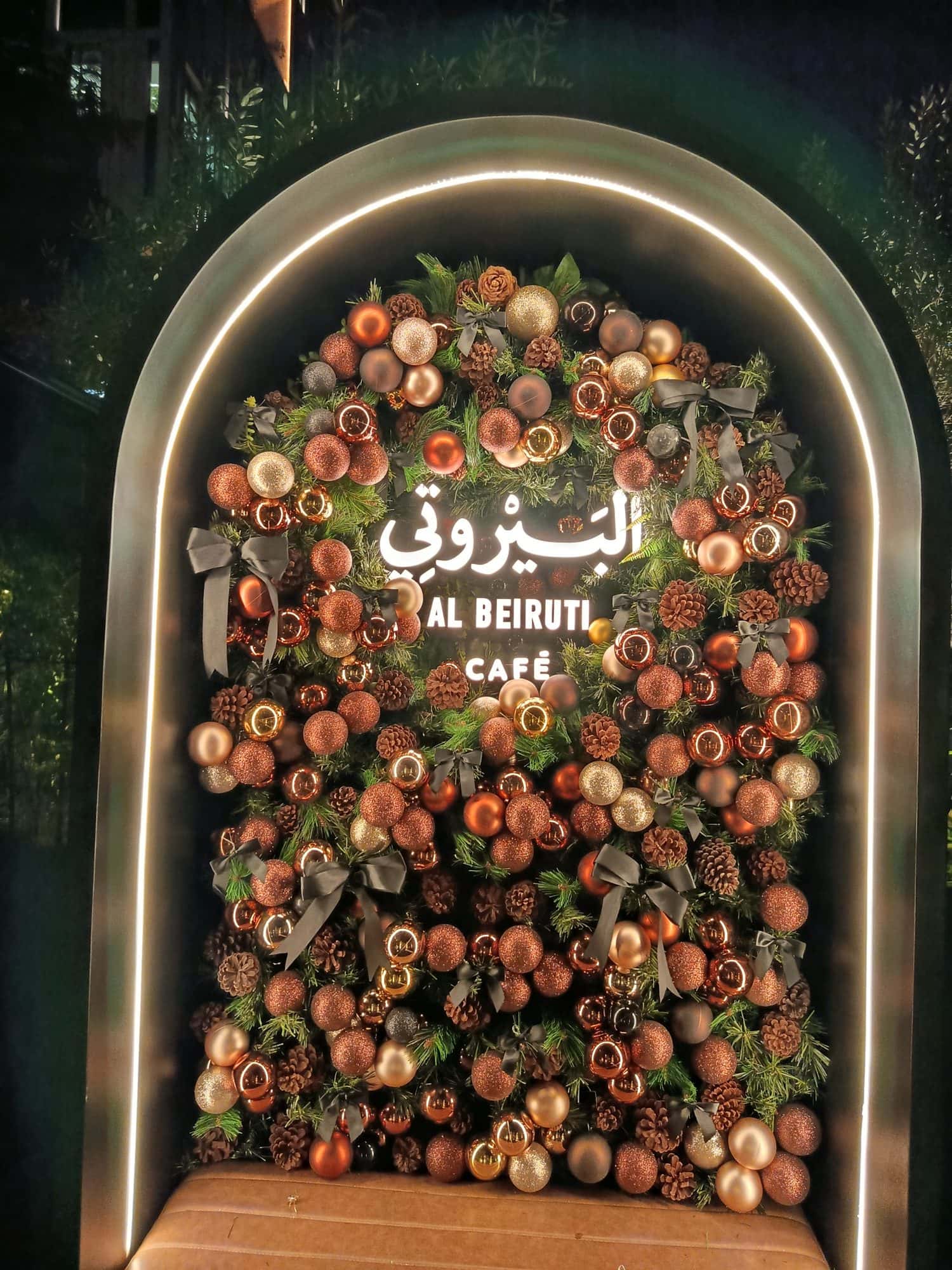 Illuminated arch filled with a dense wall of bronze and copper baubles, pine cones and grey ribbon bows, with a café’s name at the centre