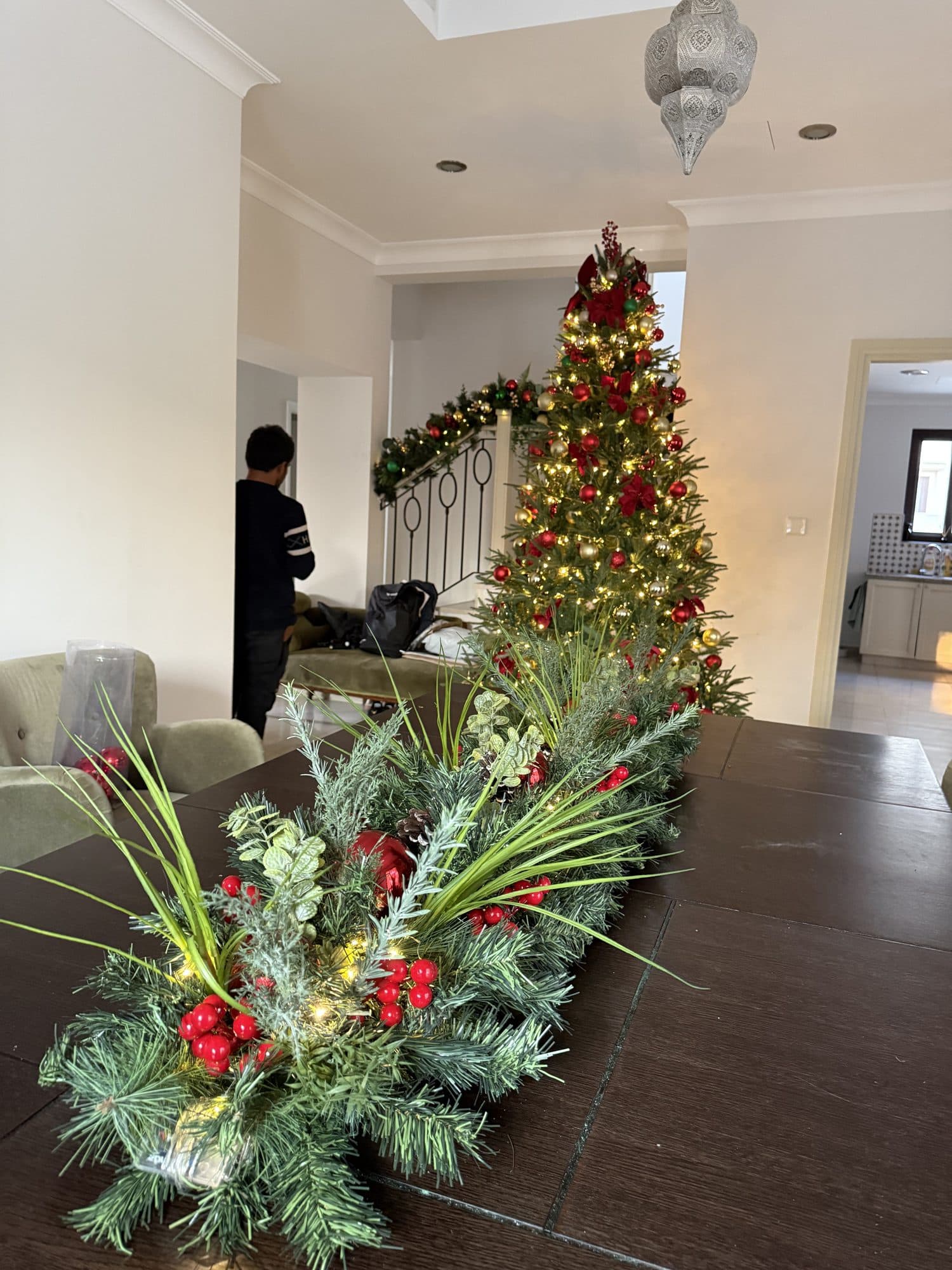 Evergreen centrepiece with red berries on a dark dining table during installation, with a lit tree and a garlanded staircase in the background
