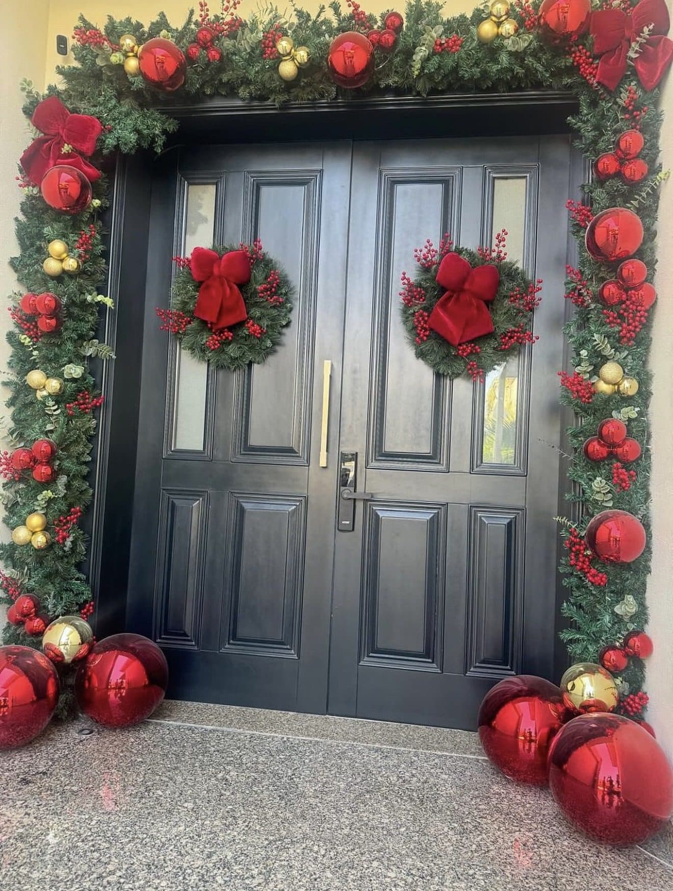 Black double front doors with twin wreaths tied in red bows, framed by a red and gold garland, with oversized red baubles on the floor either side