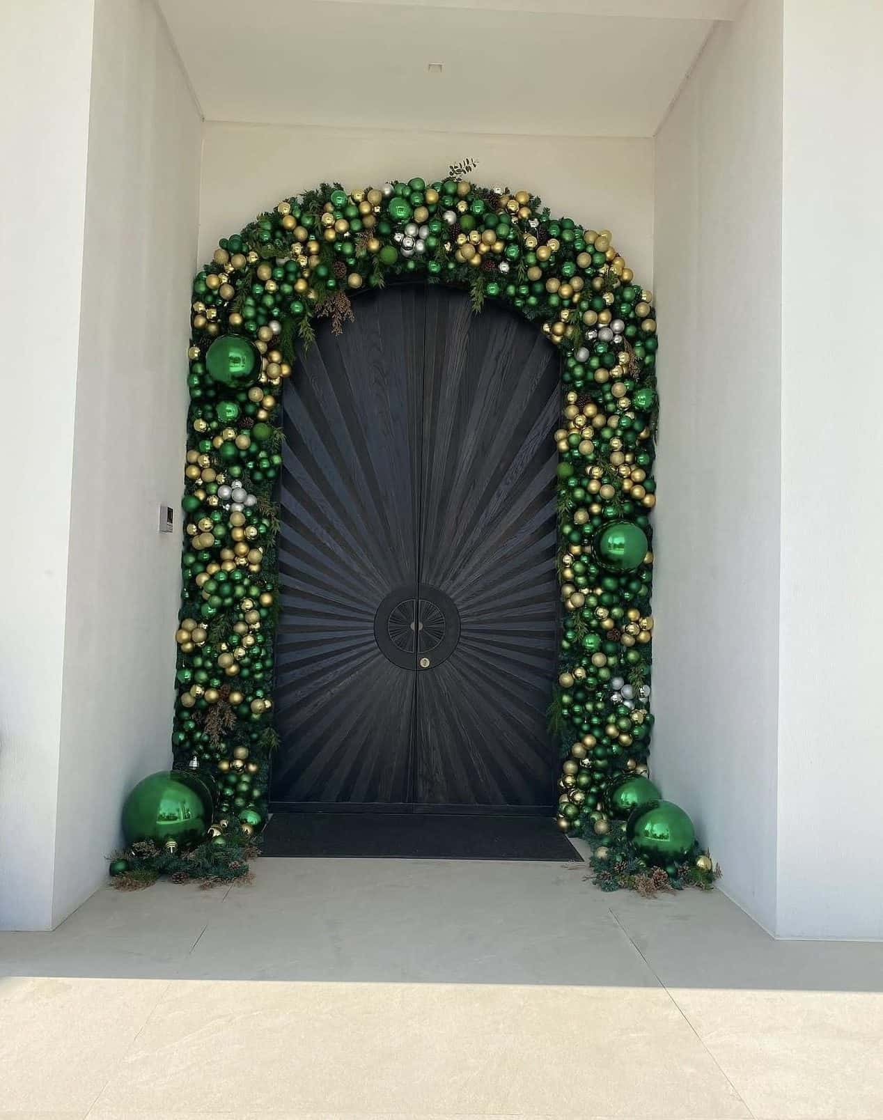 Dense arch of emerald green and gold baubles around a dark door carved in a sunburst pattern, in a white entrance