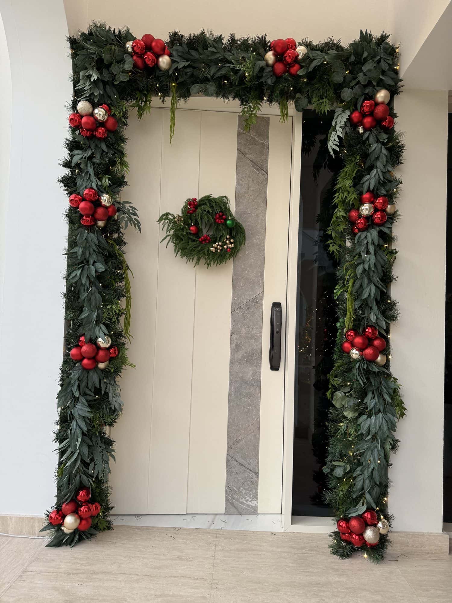Full evergreen arch with clusters of red and gold baubles framing a white modern front door with a matching wreath