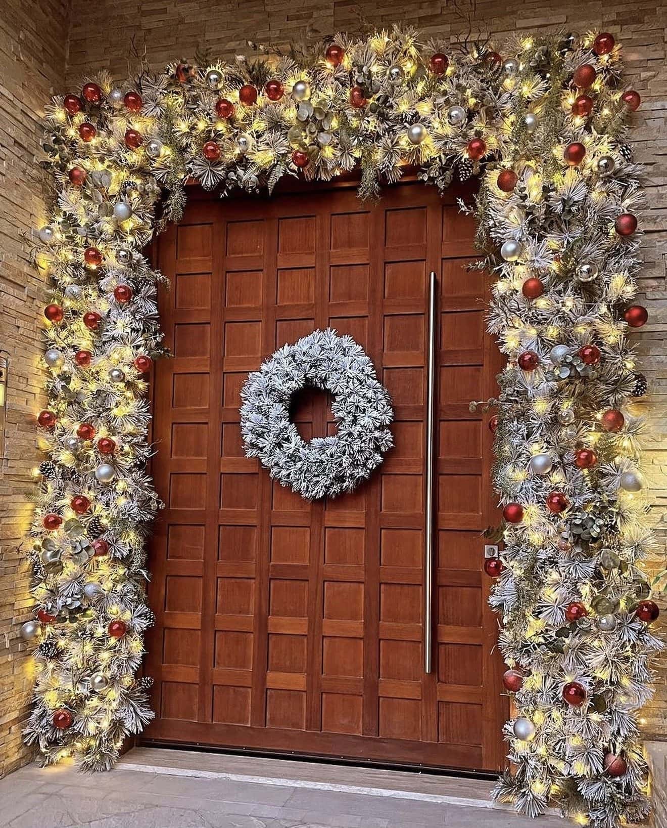 Flocked, lit garland with red and silver baubles framing a tall panelled wooden door with a frosted wreath at its centre