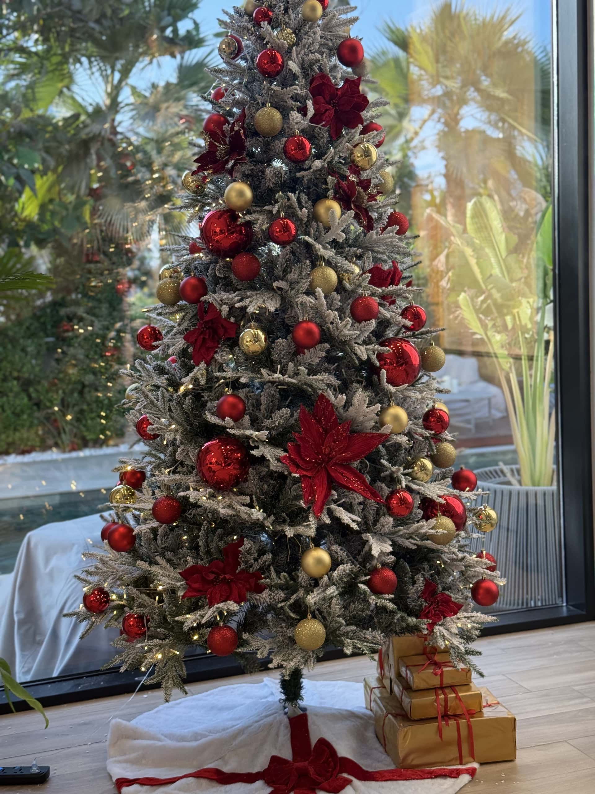 Frosted Christmas tree with red and gold baubles and red poinsettias, gift boxes and a white skirt at its base, in front of a window looking onto a pool and palms