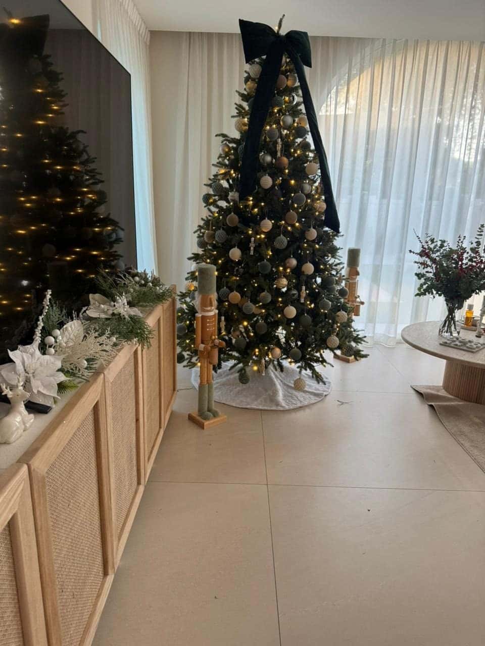 Christmas tree in green and gold baubles with an oversized dark velvet bow cascading from the top, beside a rattan sideboard with white florals