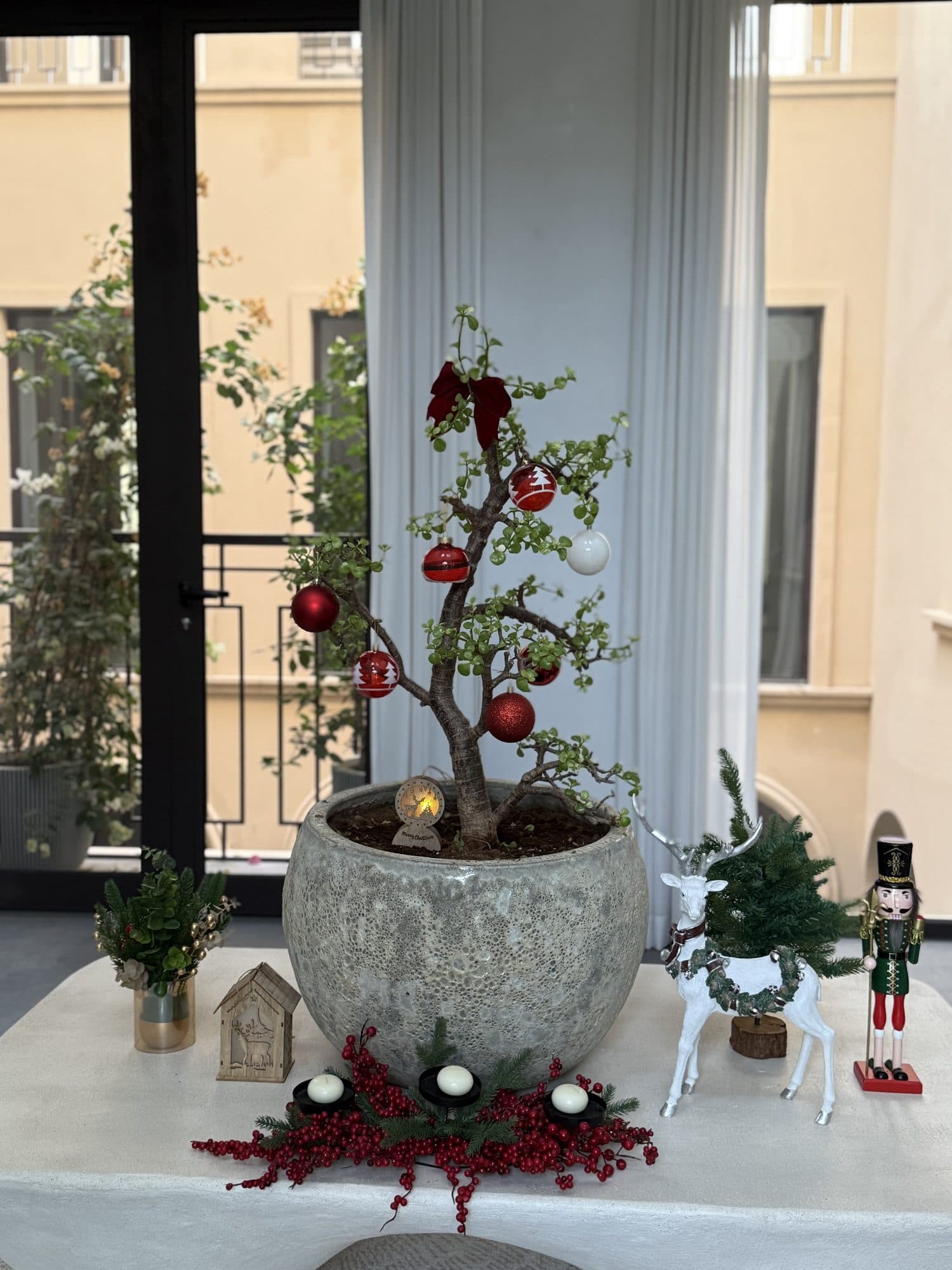 Olive tree in a large stone planter hung with red baubles and a red bow, surrounded by a nutcracker, a white reindeer, a small house and a berry garland