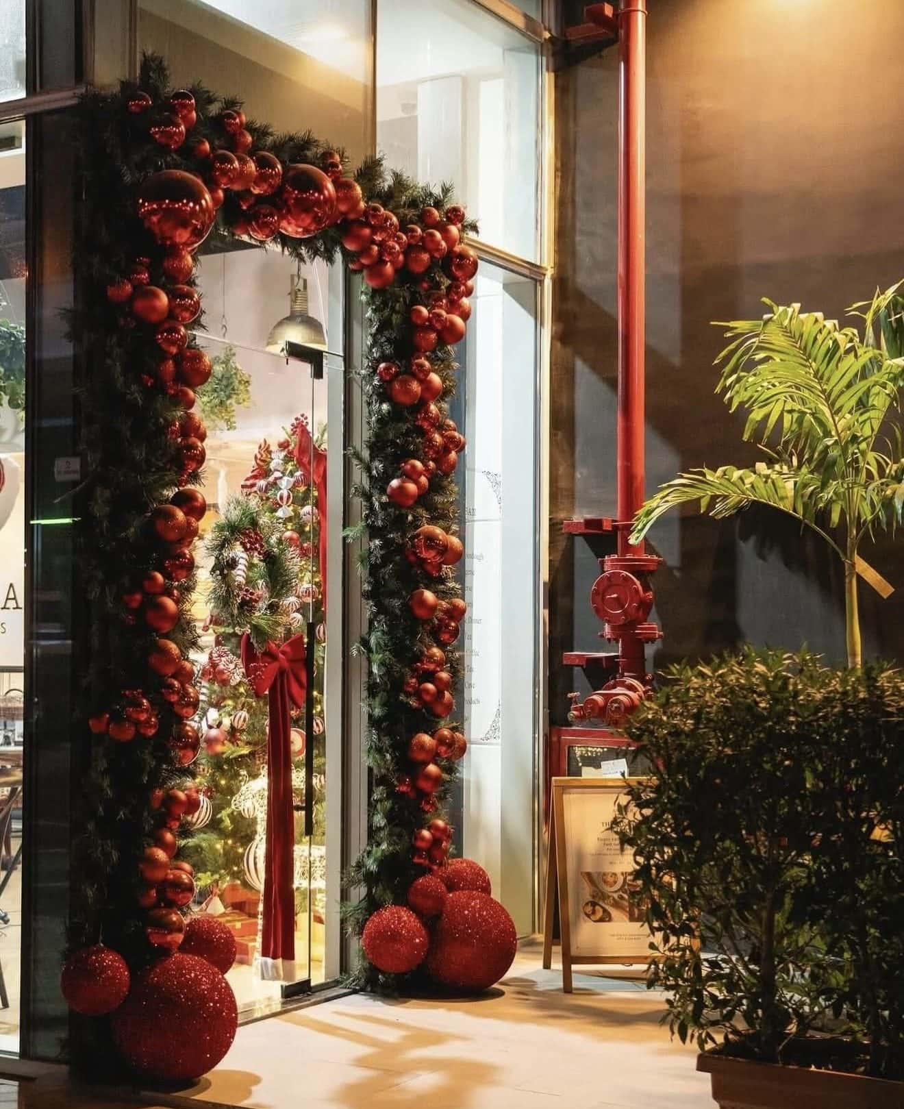 Glass shopfront in the evening framed by an arch of red baubles, with giant red baubles at the base and a decorated tree visible inside
