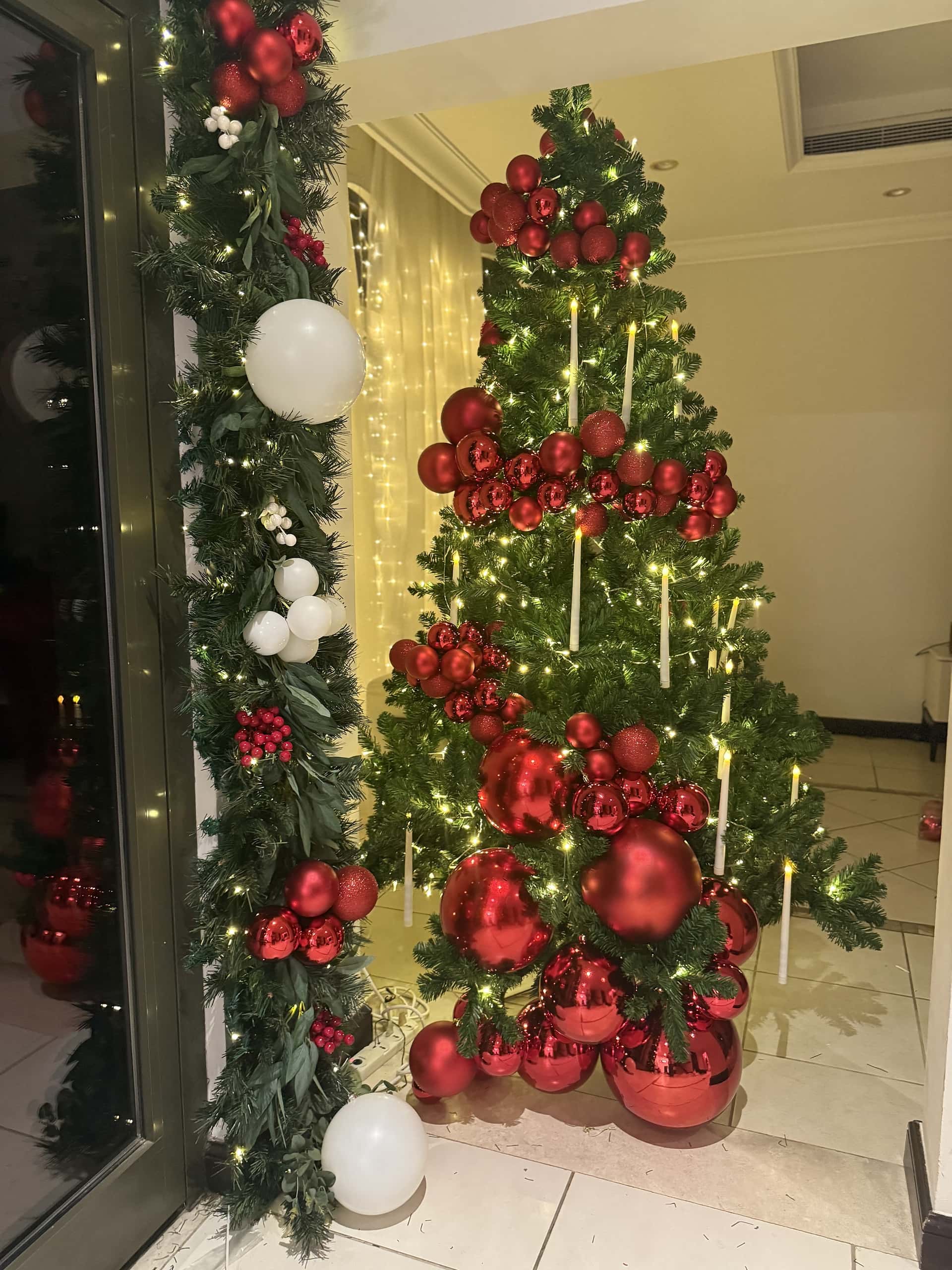Christmas tree with red baubles and candle-style lights beside a glass entrance garlanded with red baubles, white globes and berries