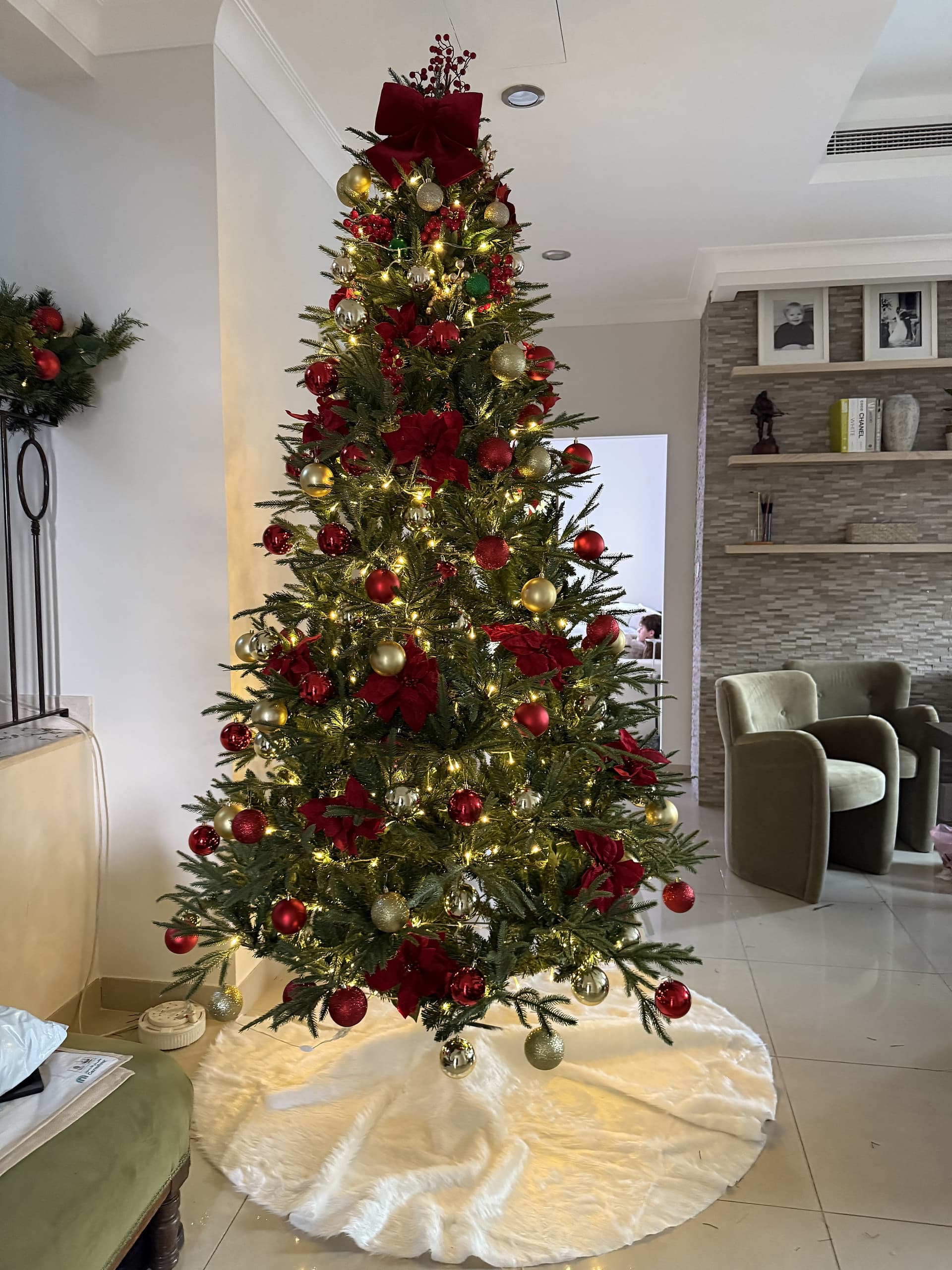 Tall Christmas tree dressed in red and gold baubles and poinsettias with a burgundy bow topper and a white faux-fur skirt in a living room