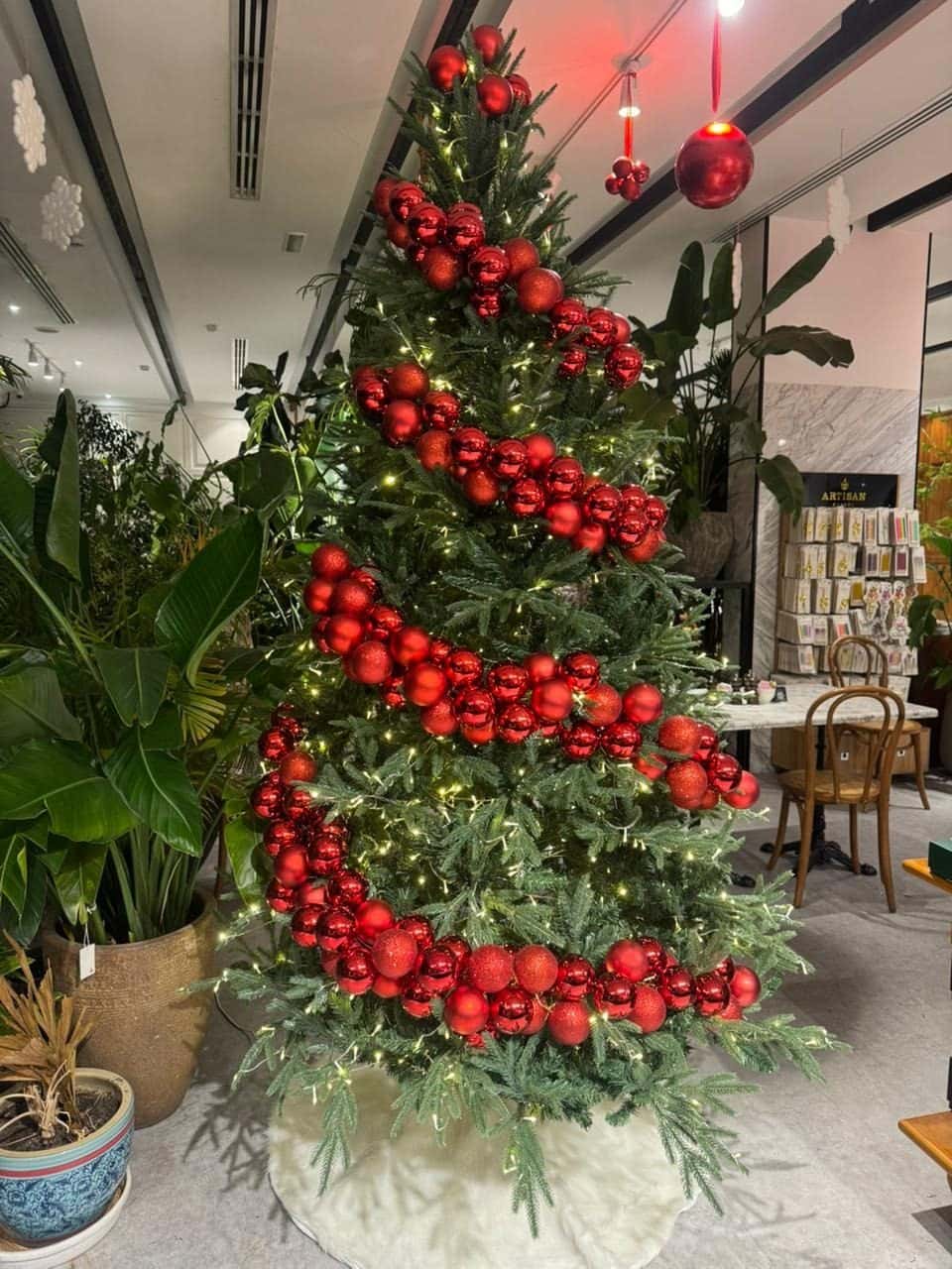 Christmas tree wrapped in spiralling garlands of red baubles among large indoor plants, with a red ornament hanging from the ceiling