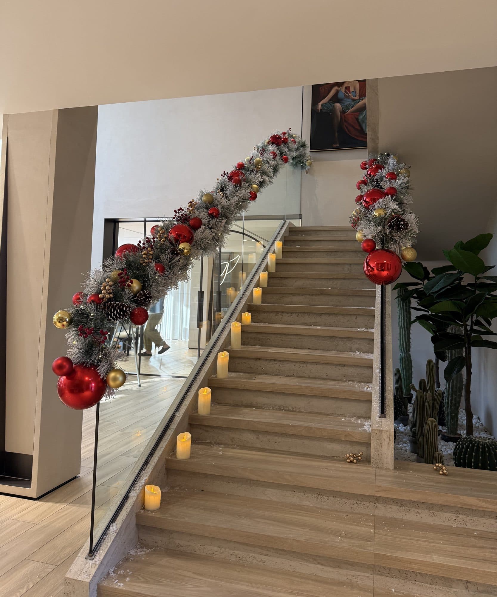 Stone staircase with a glass balustrade dressed in a frosted garland of red and gold baubles and pine cones, and candle-style lights on each step