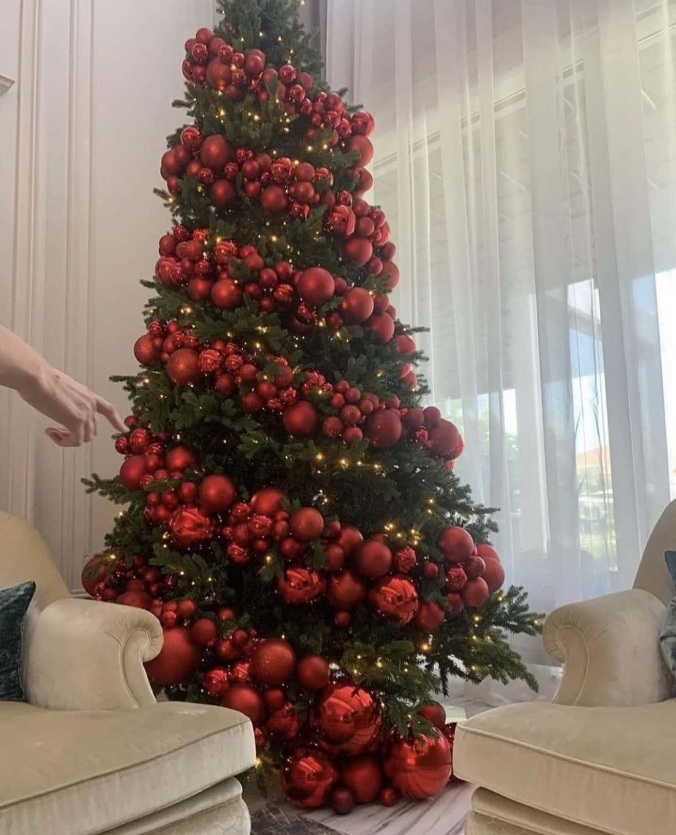 A stylist’s hand placing a red bauble on a tree dressed in red, between two cream armchairs