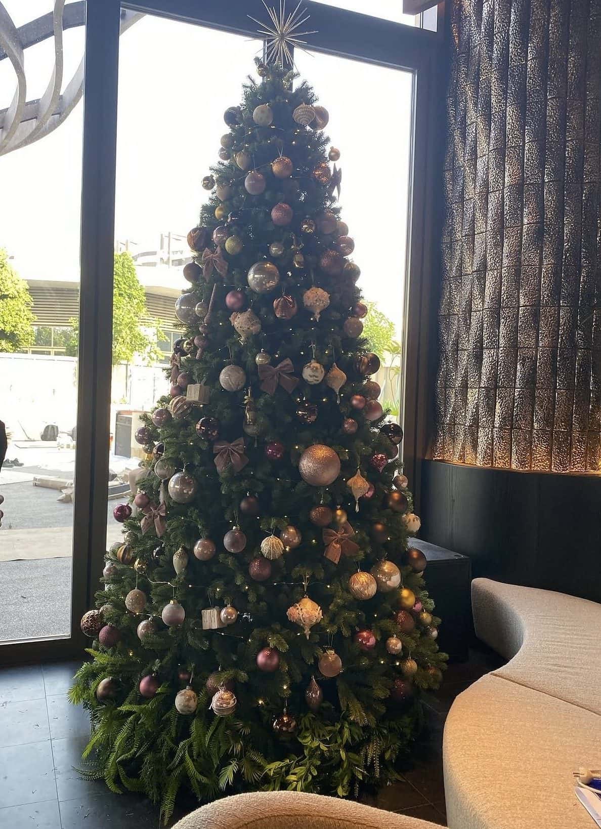 Very tall Christmas tree with bronze, blush and champagne ornaments and a star topper beside floor-to-ceiling windows in a lounge