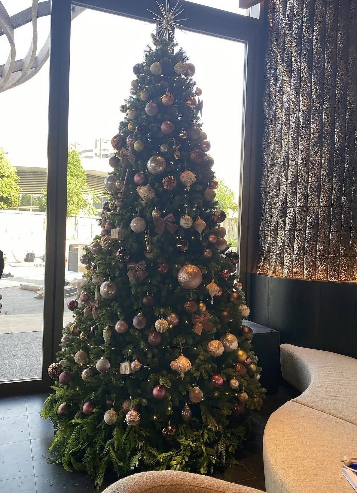 Very tall Christmas tree with bronze, blush and champagne ornaments and a star topper beside floor-to-ceiling windows in a lounge